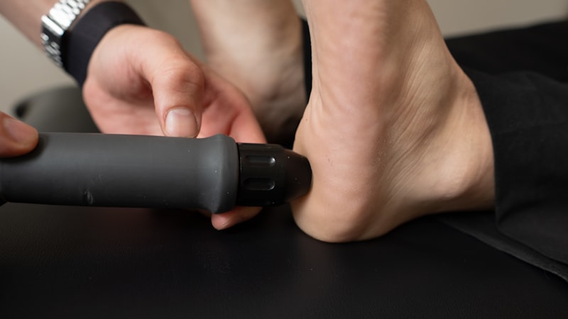 Massage gun held against the sole of a foot, showing how a deep tissue massage device is applied