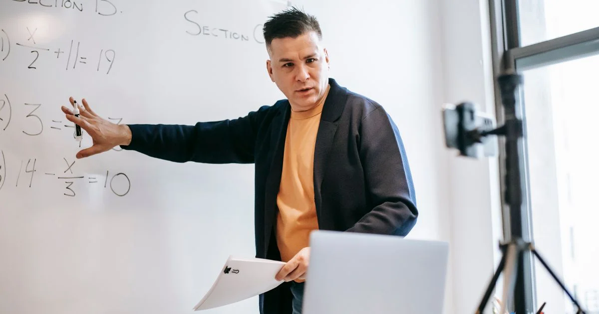 Instructor teaching a math lesson to a camera and laptop at a whiteboard, illustrating the online course vs in person comparison